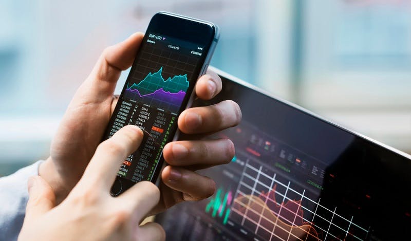 Person holding smartphone up to a laptop screen with investment graphs visible on both screens.